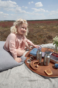 Ein Kind sitzt auf einer Wiese während eines Picknicks mit der Lässig Edelstahl-Znünibox in Blau, gefüllt mit Snacks, sowie Trinkflaschen und anderen Leckereien, ideal für Familienausflüge