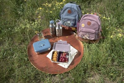 Picknick-Szene mit der Lässig Edelstahl-Znünibox in Blau, gefüllt mit frischen Snacks, zusammen mit Kinderrucksäcken auf einer Wiese, perfekt für einen Ausflug oder die Znünipause.