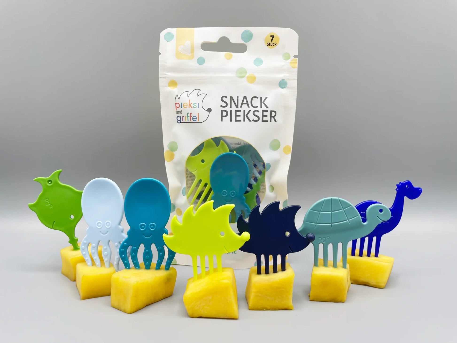 Set aus sieben bunten Gabelfiguren "Pieksi und Griffel" in verschiedenen Farben, mit Verpackung im Hintergrund und gelben Früchten auf einem Tisch arrangiert.