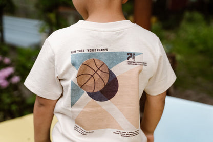 Kind trägt das T-Shirt BackPrint mit Ballsport-Print von Daily Seven und steht in einer fröhlichen Outdoor-Szene.