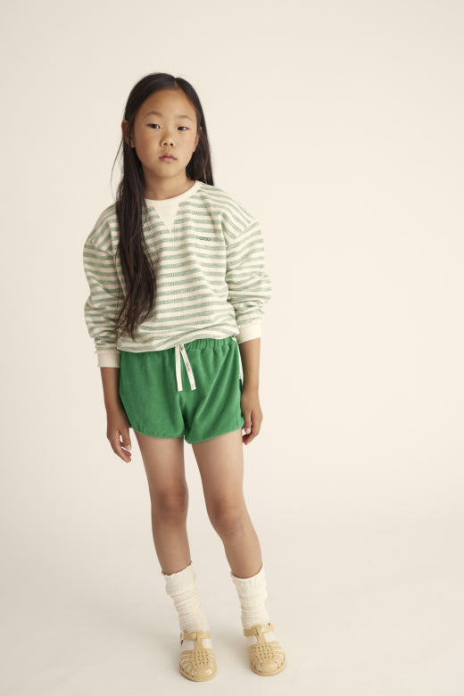 Mädchen trägt gestreiften Unisex-Kinderpullover in Grün und Creme aus nachhaltiger Bio-Baumwolle. Bequemer Schnitt, ideal für Freizeit und Spiel.